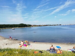 Una playa con gente tomando el sol y nadando, un barco en el agua y una costa distante con edificios.