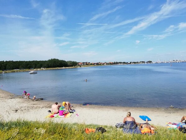 Una playa con gente tomando el sol y nadando, un barco en el agua y una costa distante con edificios.