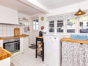 Una cocina con armarios blancos, encimera de madera, horno negro, refrigerador y ventana con cortinas florales.