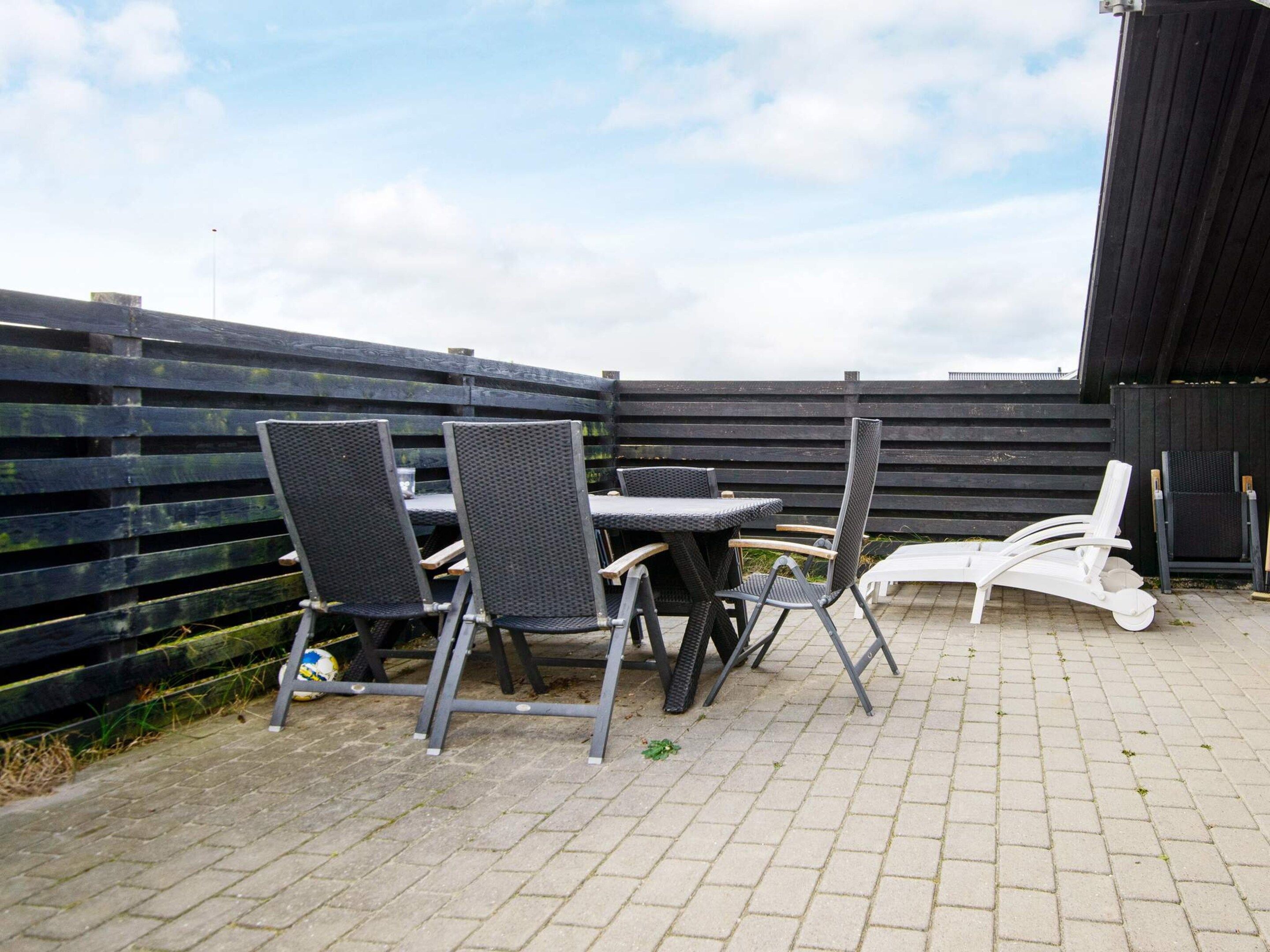 An outdoor patio area with a table and chairs, a white lounge chair, and a wooden fence.