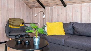 A room with a wooden ceiling, a grey sofa, a small round table with a plant, a lamp, and a chair with a blanket.