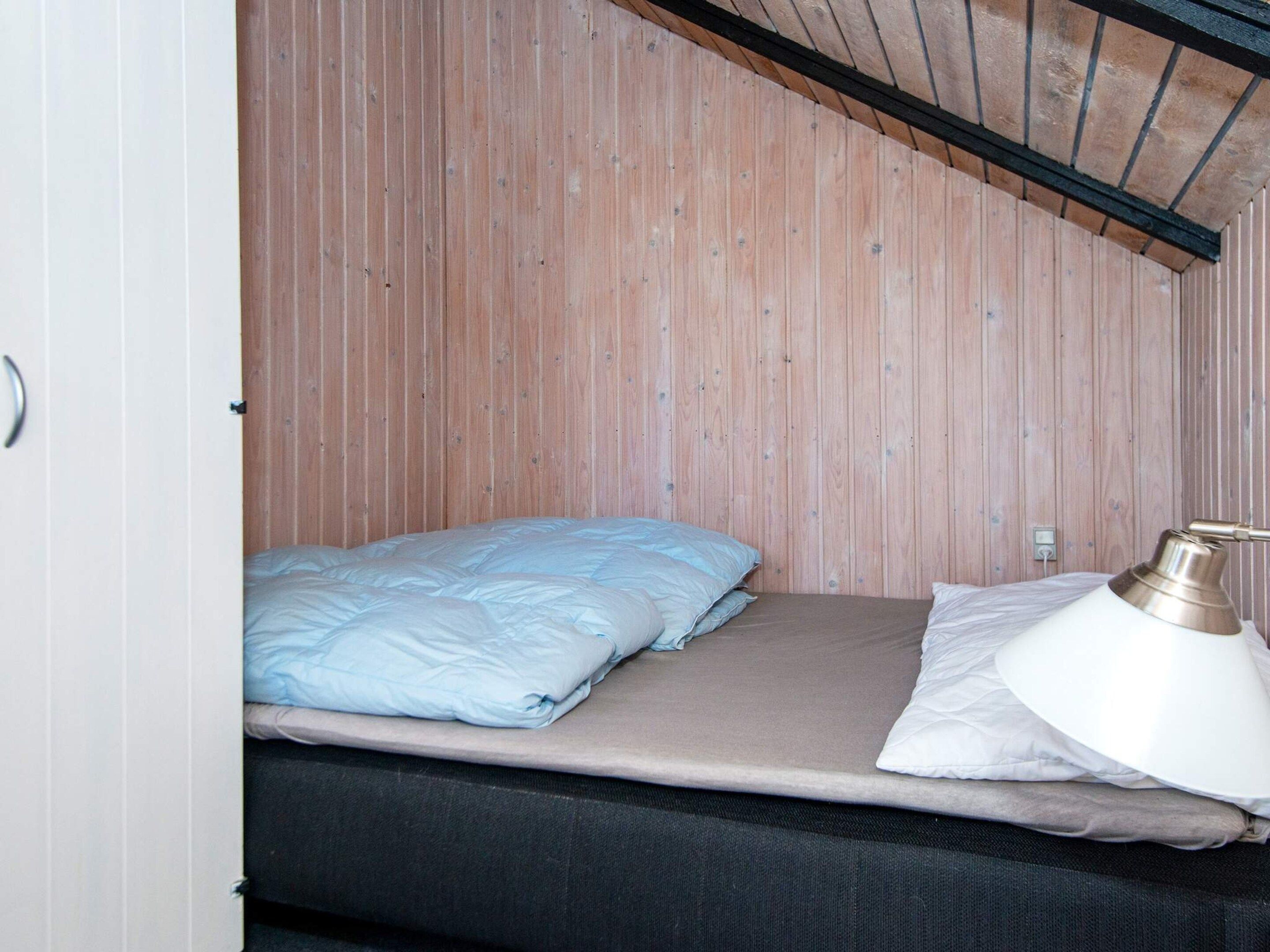 Una cama individual con un almohadón blanco y un colchón color beige, una lámpara blanca en la mesita de noche y un techo de listones de madera.