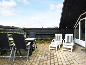 An outdoor patio with a table and chairs, a wooden fence, and a building with a window. - 4 Person Holiday Home in Hvide Sande (Hvide Sande)