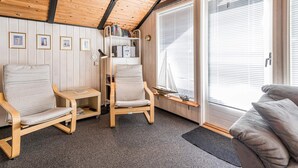 Une pièce chaleureuse aux murs et au plafond en bois, comprenant deux chaises en bois, une petite table, une bibliothèque et une grande fenêtre avec des stores.