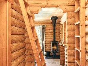 A log cabin interior with a central wood-burning stove, wooden walls, and a ladder.