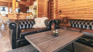L’intérieur d’une cabane en rondins, avec un canapé en cuir, une table en bois et une cuisine à l’arrière-plan.