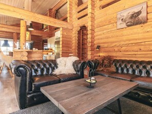 L’intérieur d’une cabane en rondins, avec un canapé en cuir, une table en bois et une cuisine à l’arrière-plan.