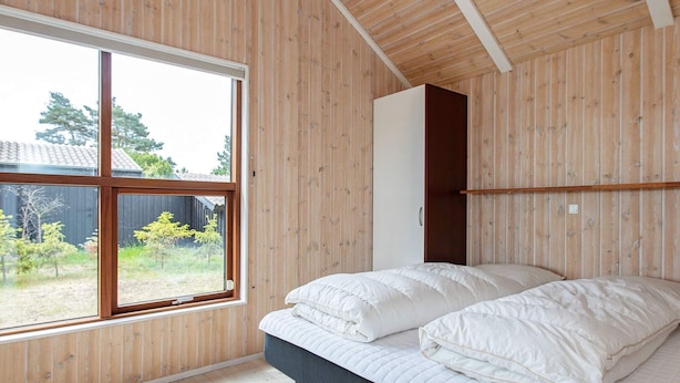 A bedroom with a large window, wooden walls, a bed with white bedding, and a wardrobe. - Holiday Home in Gedser (Gedser)
