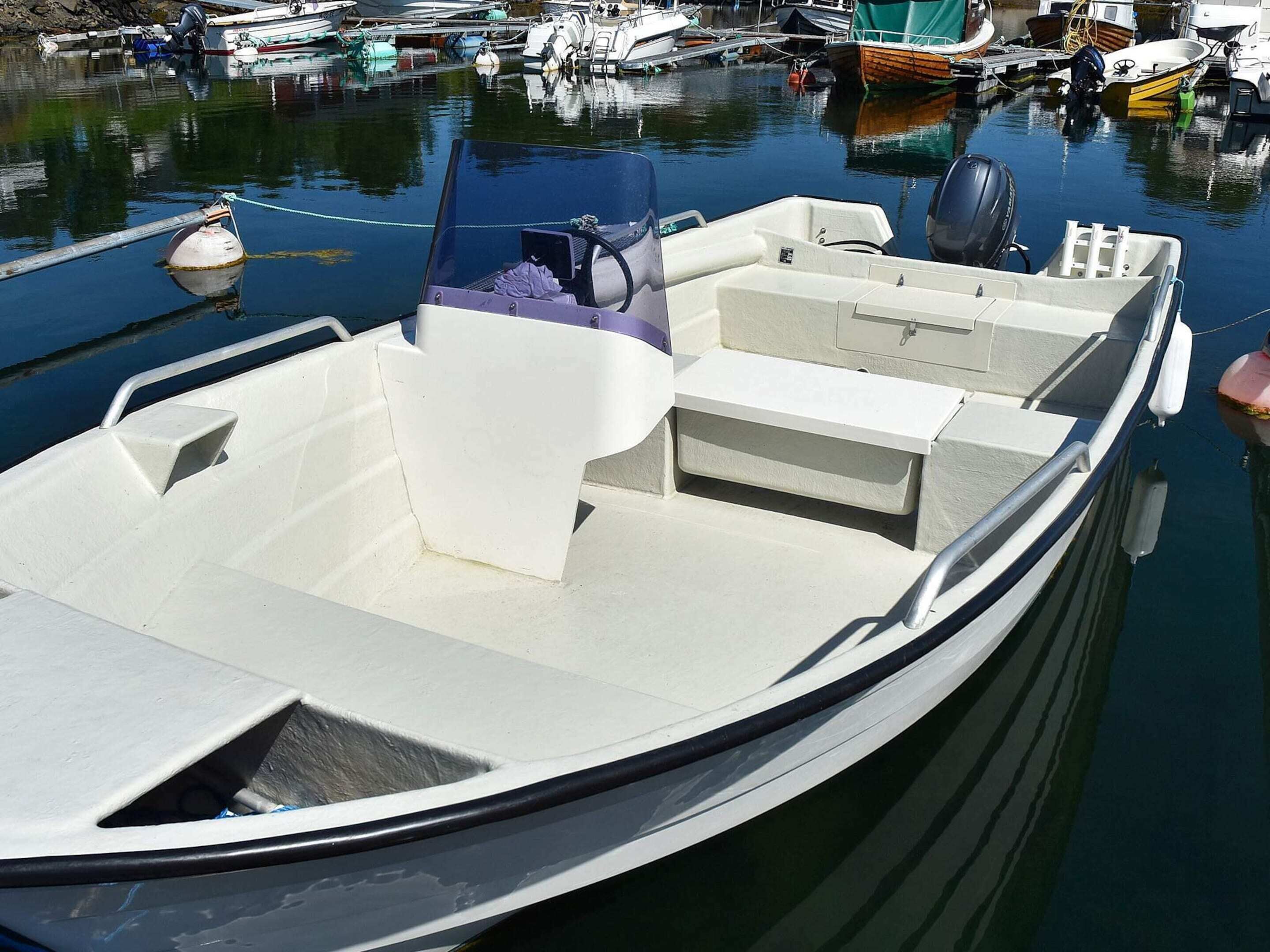 Ein kleines Motorboot liegt in einer Marina festgemacht, mit hellem Innenraum, einem Motor am Heck und einem Wakeboard auf dem Deck.