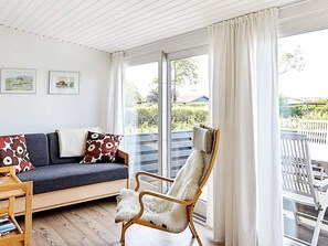 A cozy room with a sofa, a chair, and a table. There are large windows with curtains, and a balcony with outdoor furniture.