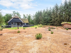 A rustic cabin with a thatched roof, surrounded by a forested area with a dirt ground. - 4 Star Holiday Home in Norre Nebel (Norre Nebel)
