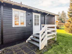 A dark wooden house with a white front door, a small porch with steps, and a clear view of the surrounding garden. - 6 Person Holiday Home in Vaeggerlose (Vaeggerlose)