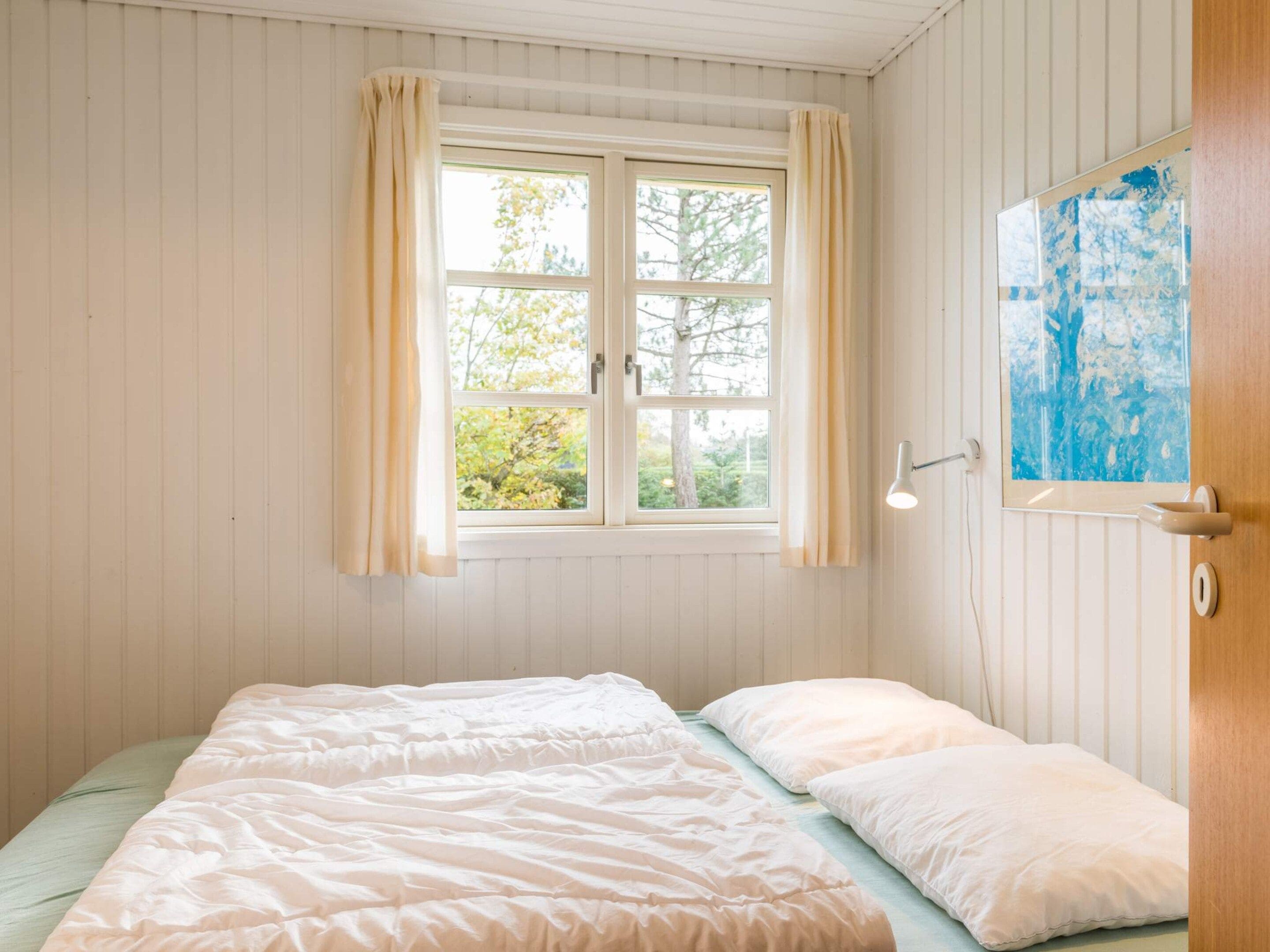 A room with a bed, a window with curtains, a wall-mounted lamp, and a door with a painting.