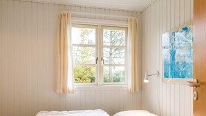 Une pièce avec un lit, une fenêtre avec des rideaux, une lampe fixée au mur et une porte ornée d’un tableau.