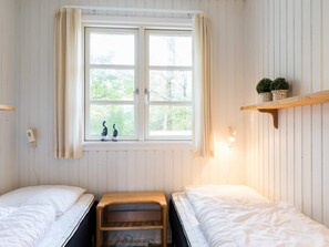 A room with two beds, a window with curtains, a shelf with plants, and a wall-mounted light.