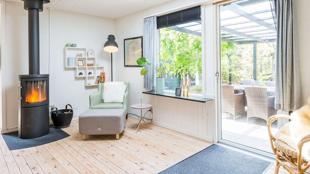 A cozy room with a wood-burning stove, a comfortable seating area with a sofa and chairs, a small table, and a large window with a view of greenery. - 2 Person Holiday Home in Henne (Henne)