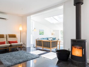 Een moderne woonkamer met een zwart houtgestookte kachel, een bank met geruite kussens en uitzicht op een tuin door dakramen.