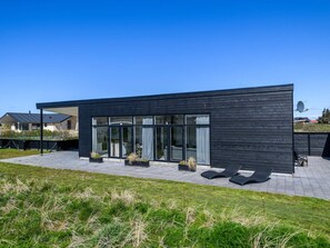 A modern house with large glass windows, a flat roof, and a paved patio area with outdoor furniture.