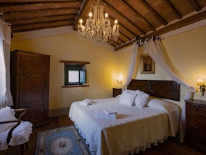 Quarto com uma cama grande, vigas de madeira no teto, um candelabro, uma janela pequena e uma mesa de cabeceira com uma luminária.
