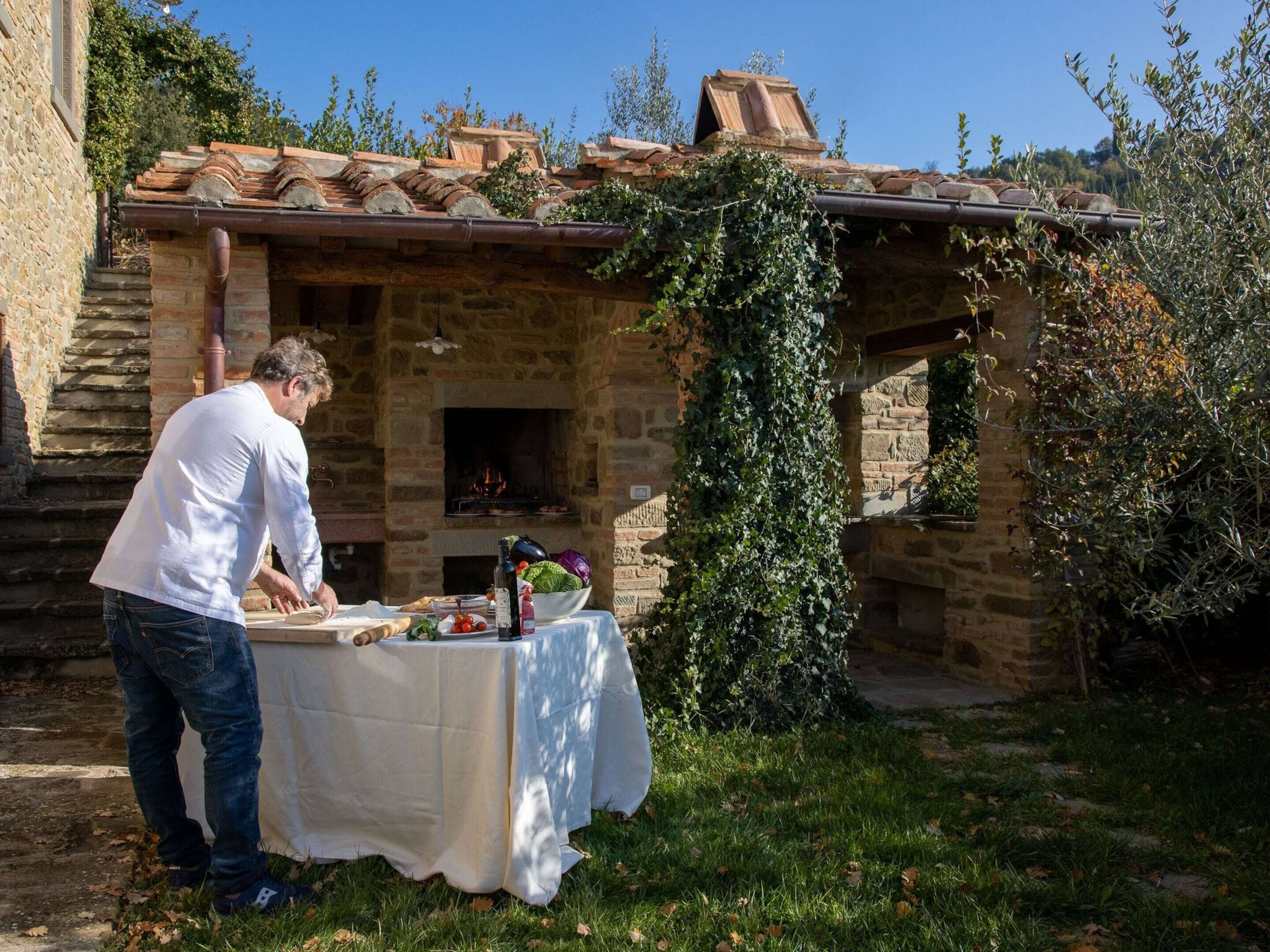 A man preparing food outdoors next to a stone building with a brick roof and a fire pit.