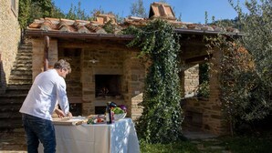 A man preparing food outdoors next to a stone building with a brick roof and a fire pit. - Holiday Home in Cortona (Cortona)