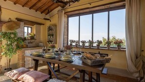 A rustic dining area with a wooden table set for a meal, a large window with a view, and potted plants on the windowsill. - Holiday Home in Cortona (Cortona)
