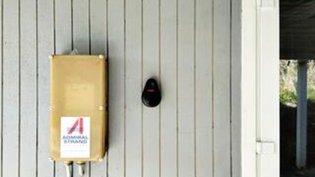 Una pared blanca con un dispositivo de seguridad negro y una caja eléctrica marrón con una etiqueta.
