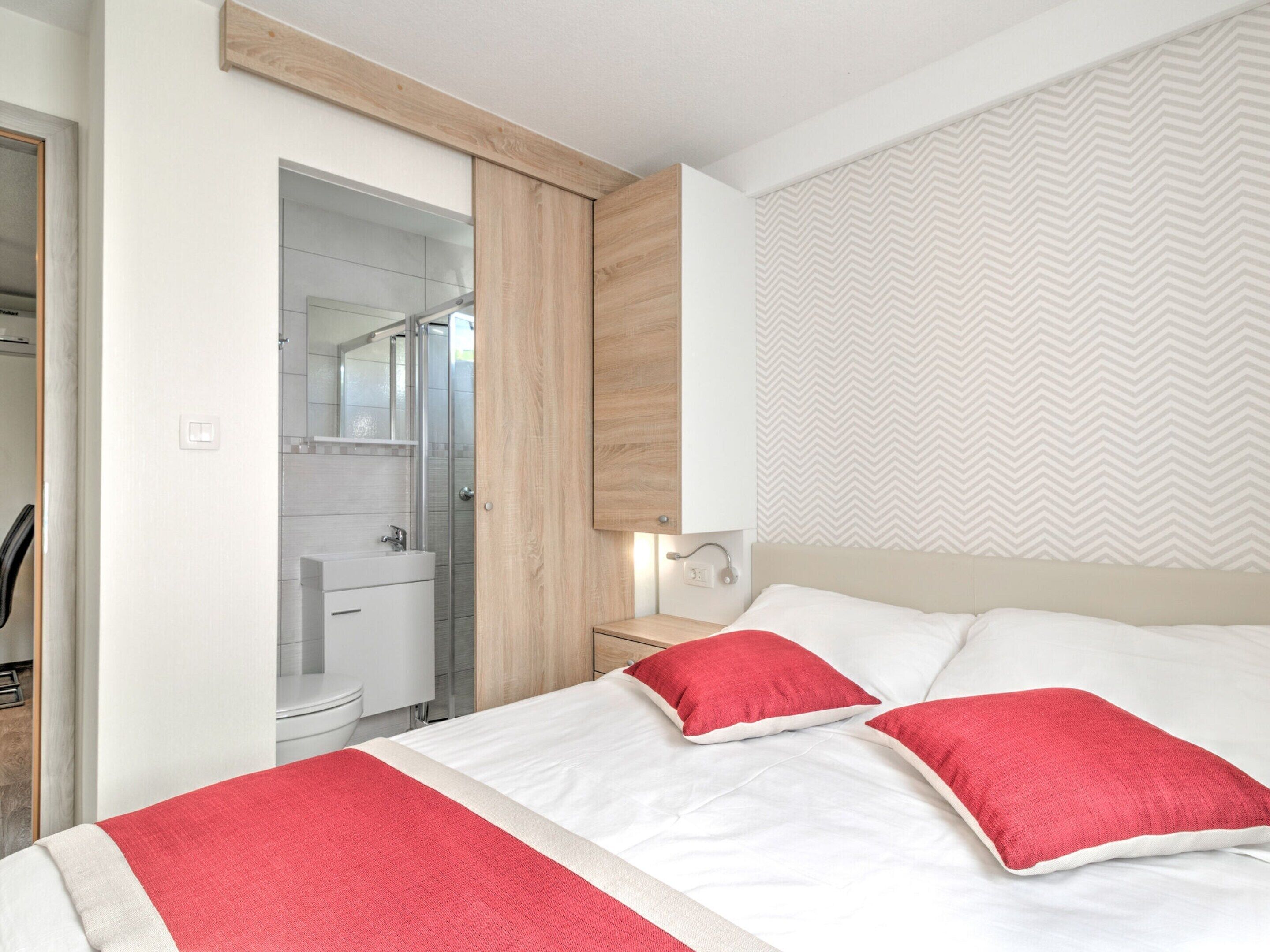 A bedroom with a bed, two red pillows, a wooden headboard, a nightstand, and a bathroom with a shower and toilet.