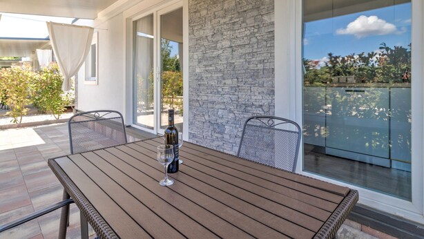 Une table en bois, avec une bouteille de vin et un verre, sur un balcon doté d’un mur en pierre et de portes vitrées.