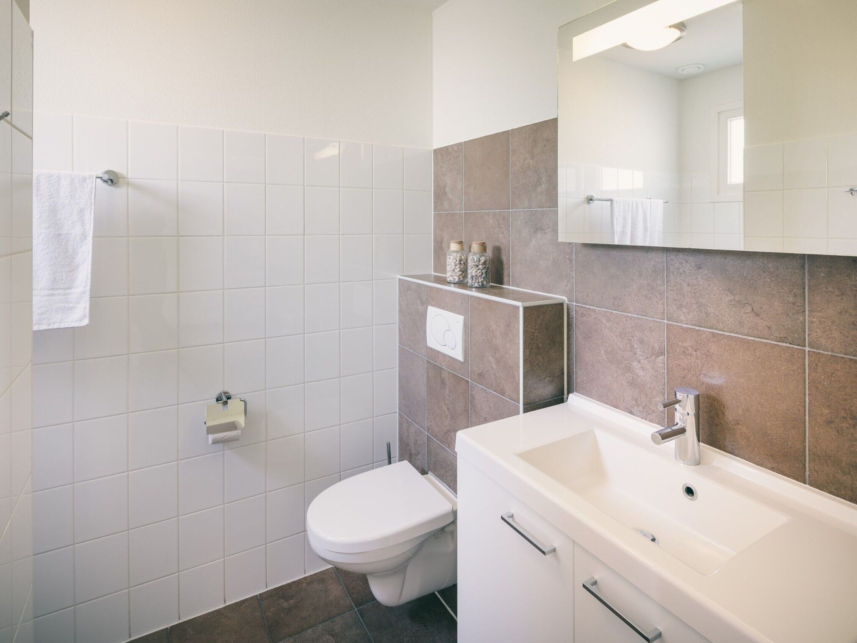 A modern bathroom with white and beige tiles, a white toilet, a sink with a mirror, and a shower area.
