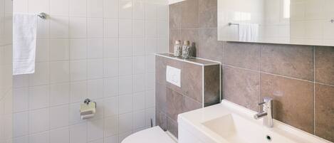 A modern bathroom with white and beige tiles, a white toilet, a sink with a mirror, and a shower area.