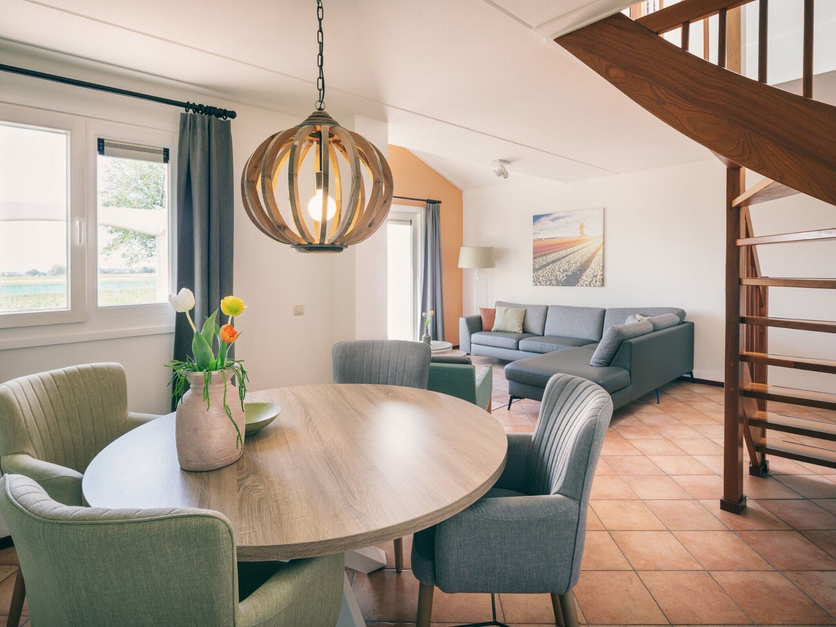 A modern living room with a round wooden table, grey upholstered chairs, a grey sofa, and a wooden staircase.