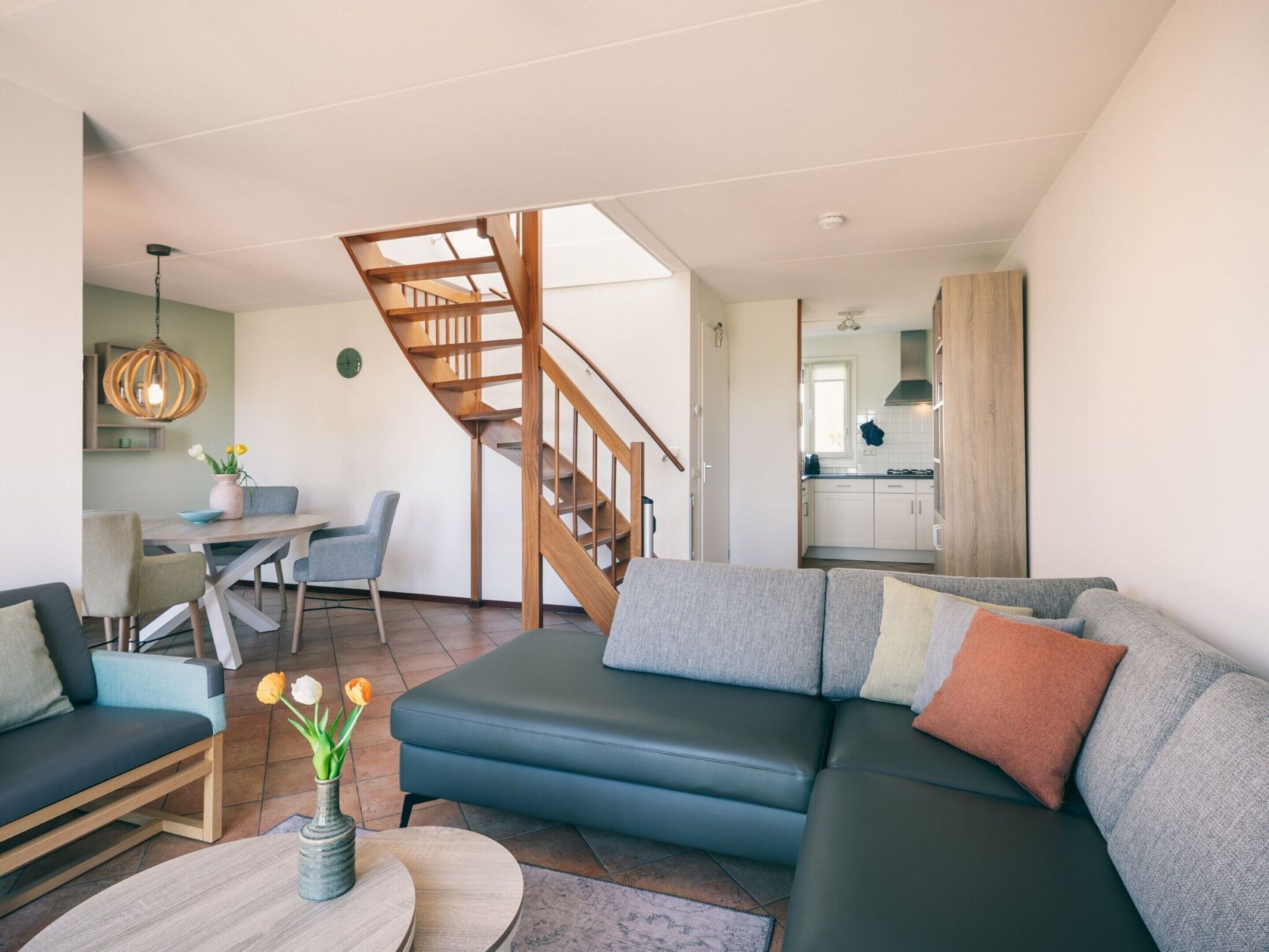 Ein modernes Wohnzimmer mit einer Couch, einem Couchtisch und einem Essbereich mit Stühlen. Eine Holztreppe führt auf eine obere Ebene.