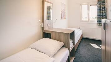 A hotel room with two beds, a wooden desk, a mirror, a radiator, and a window with curtains.