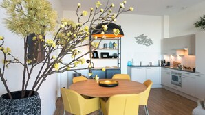 Une cuisine moderne avec des meubles blancs, une table à manger en bois et des chaises jaunes, ainsi qu’une plante décorative aux fleurs jaunes.