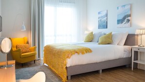 Une chambre d’hôtel moderne avec un grand lit, un fauteuil jaune, une table de chevet avec une lampe et une grande fenêtre ornée de rideaux légers.
