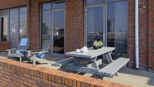 Une terrasse avec une table et des chaises, un mur en briques et de grandes fenêtres.