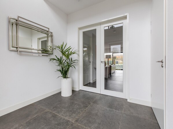 Un pasillo moderno con una planta en maceta, un espejo y una puerta que da a una habitación con cama y lámpara.