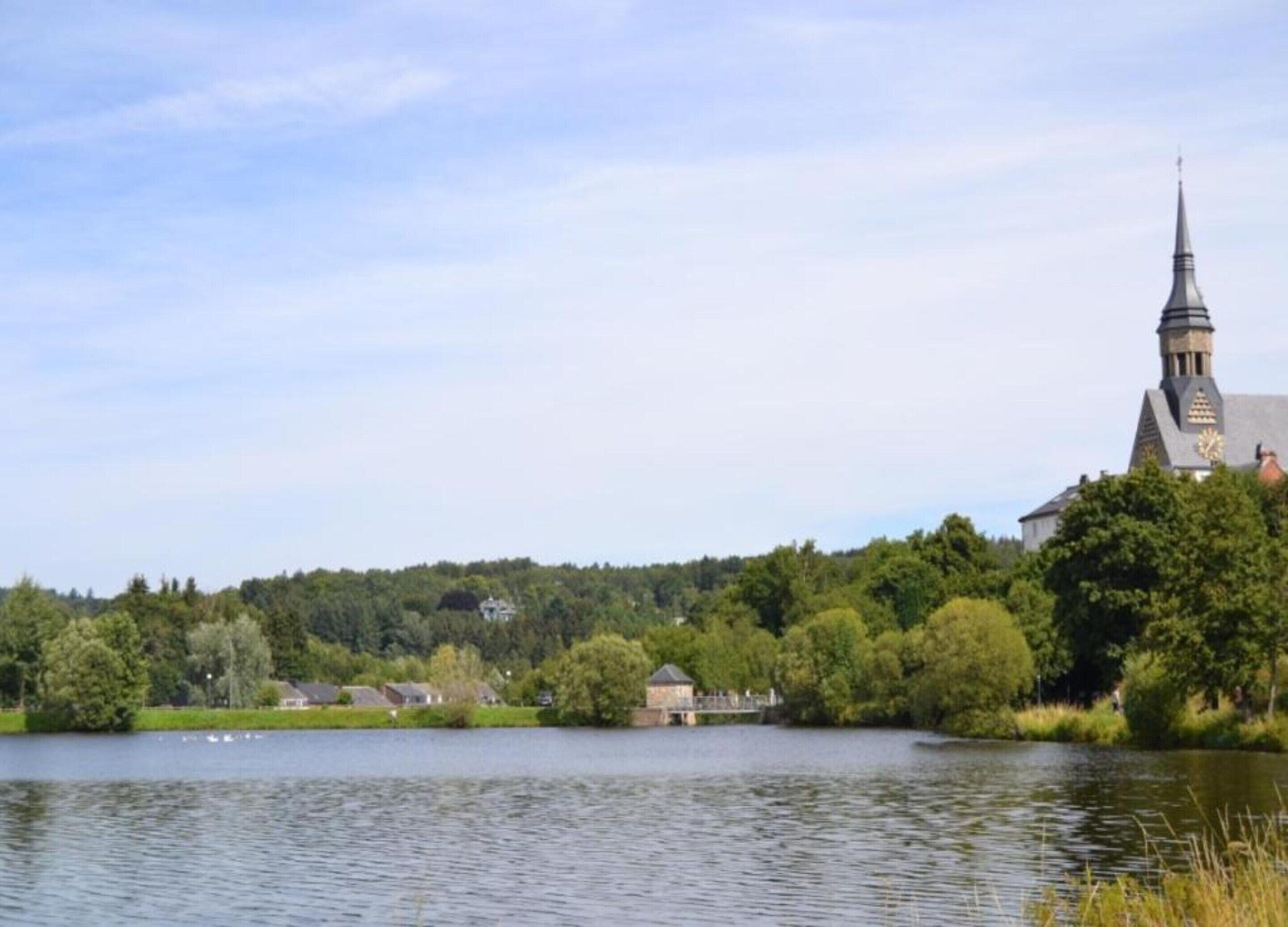A church with a tall steeple and a clock, surrounded by trees and a body of water.