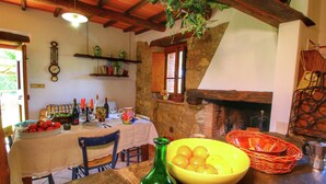Un coin repas rustique avec une cheminée en pierre, des poutres en bois et une table dressée avec un panier de fruits, une bouteille verte et divers plats.