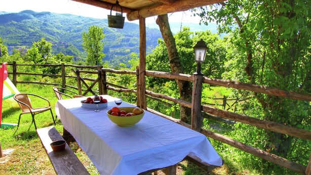 Une table en bois recouverte d’une nappe blanche, un saladier rempli de fruits et un verre à vin posés sur un banc en bois.