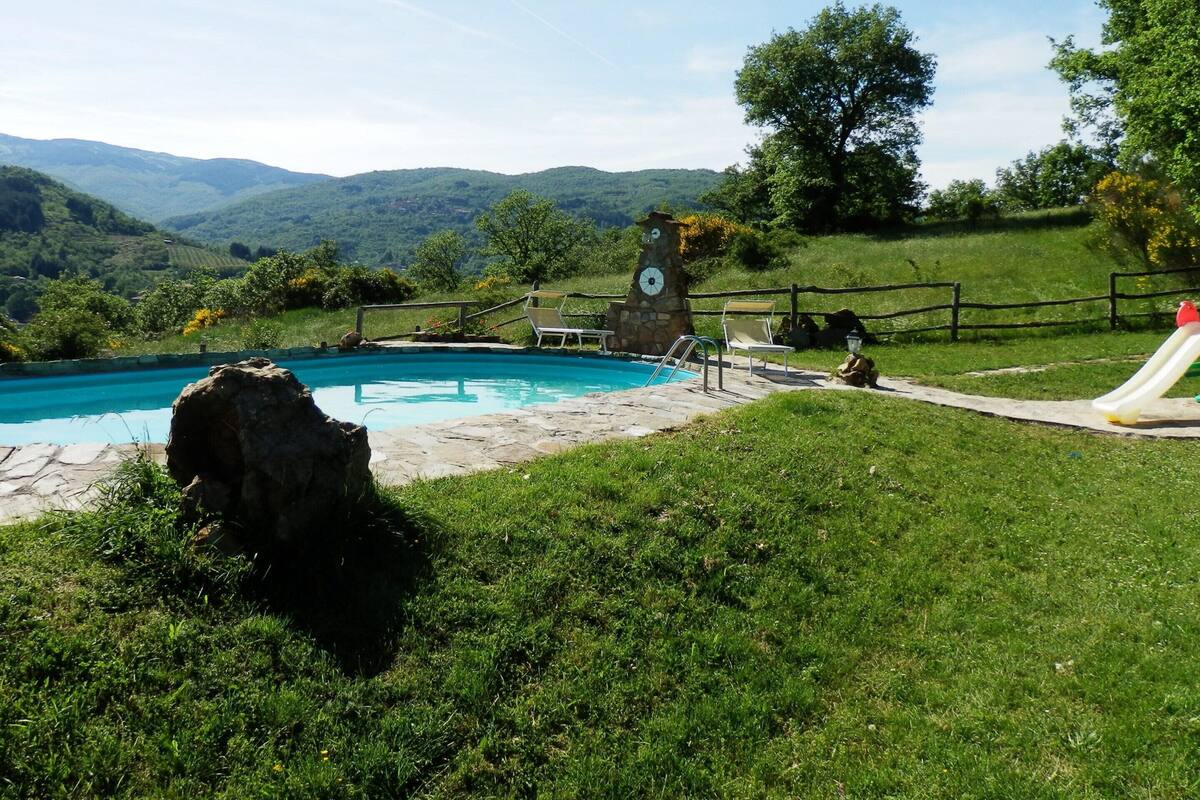 A swimming pool with a stone clock tower, a slide, and a grassy area with mountains in the background.