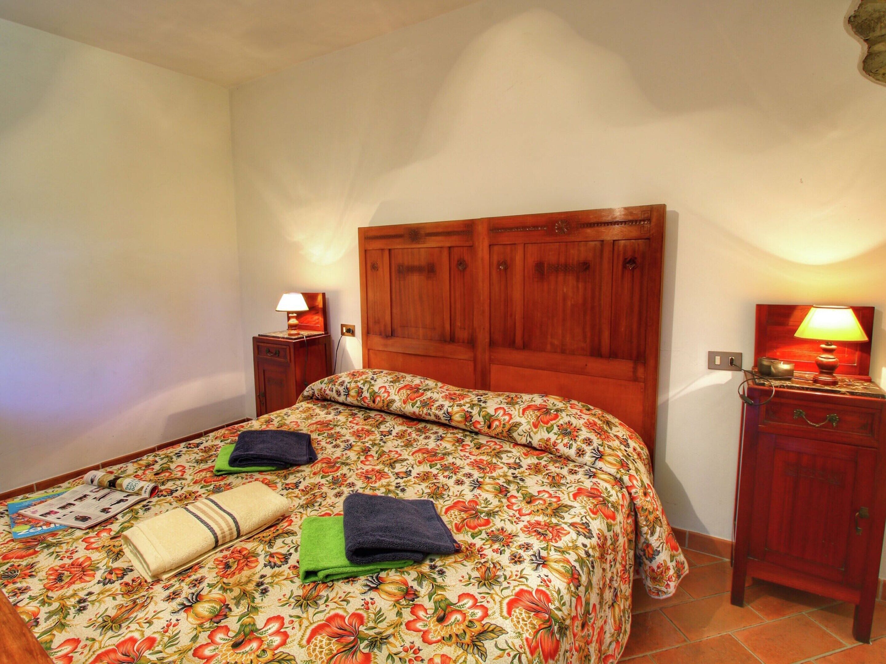A bedroom with a wooden headboard, a floral bedspread, two bedside tables with lamps, and a neatly made bed with towels.