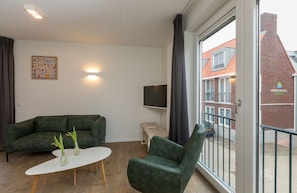 A modern apartment with a sofa, a small table, a TV, and a balcony with a view of a red brick building. - Stylish Beach Getaway (Zoutelande)
