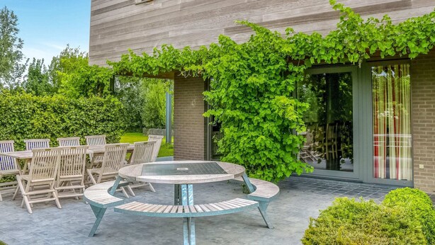 A modern house with a patio area featuring a round table and chairs, surrounded by greenery and a wooden fence. - Holiday Home in Gijverinkhove Near Plopsaland (Alveringem)