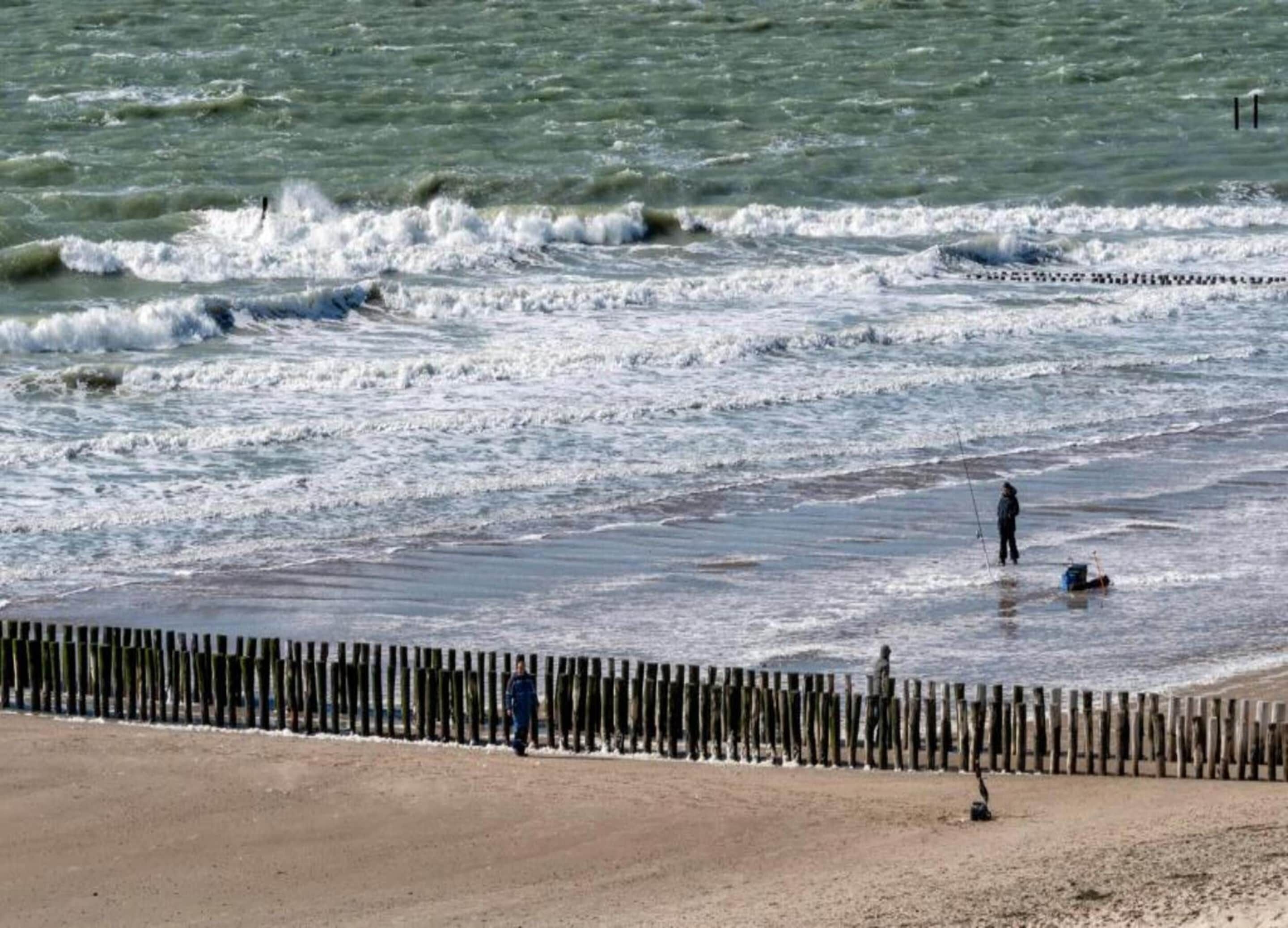 A beach with a wooden fence, waves, and a person fishing.