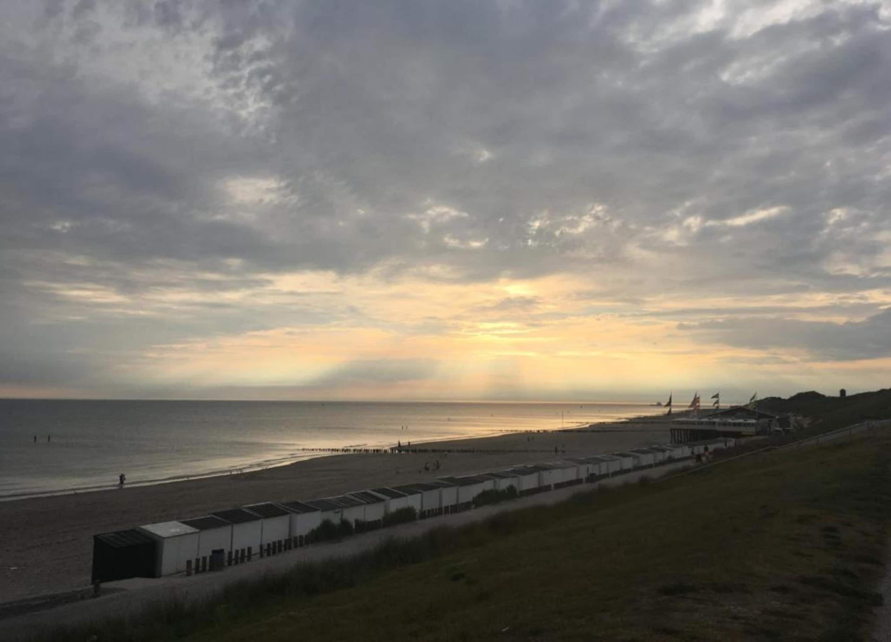 Una playa con una hilera de edificios blancos a lo largo de la orilla, un cielo nublado y el sol poniéndose o saliendo cerca del horizonte.