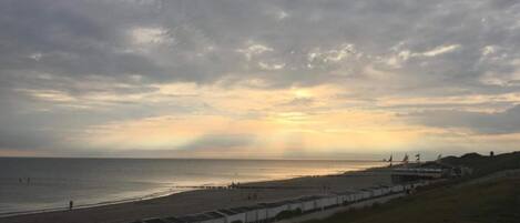 Una playa con una hilera de edificios blancos a lo largo de la orilla, un cielo nublado y el sol poniéndose o saliendo cerca del horizonte.