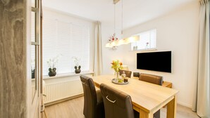 A dining area with a wooden table, brown chairs, a TV mounted on the wall, and potted plants on the windowsill. - Holiday Home in Kilder Near Bergherbos (Kilder)