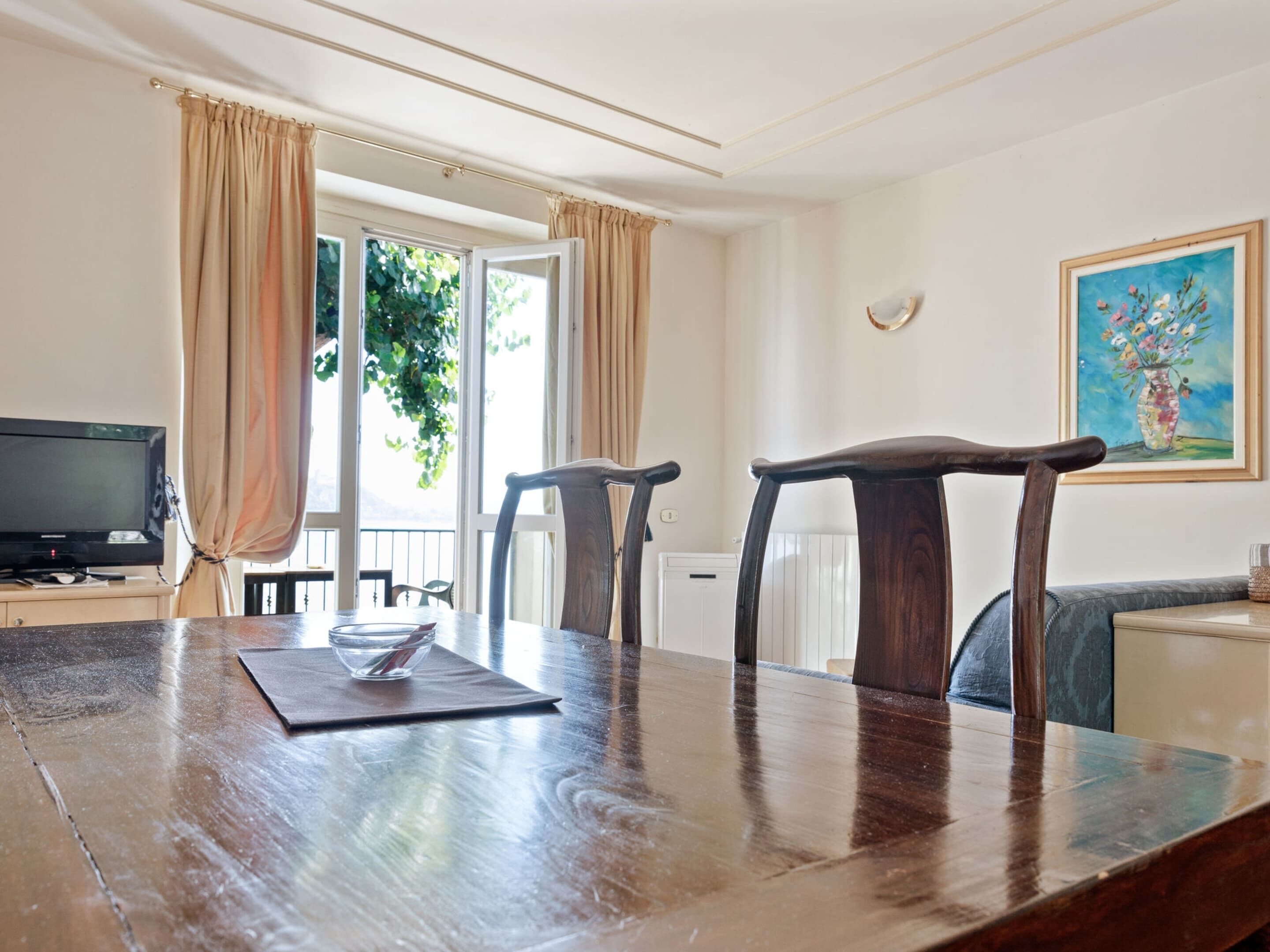A dining area with a wooden table, two chairs, a television, and a painting on the wall.